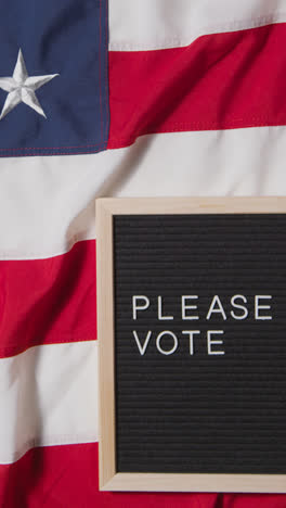 Vertical-Video-Of-Sign-Reading-Please-Vote-Lying-On-US-Stars-And-Stripes-Flag-For-American-Election-2024-1